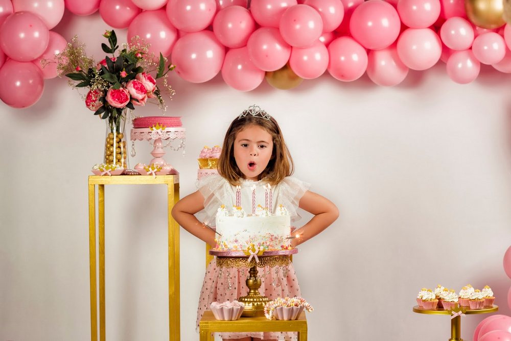 Birthday girl in a tiara blowing out candles under a pink balloon arch